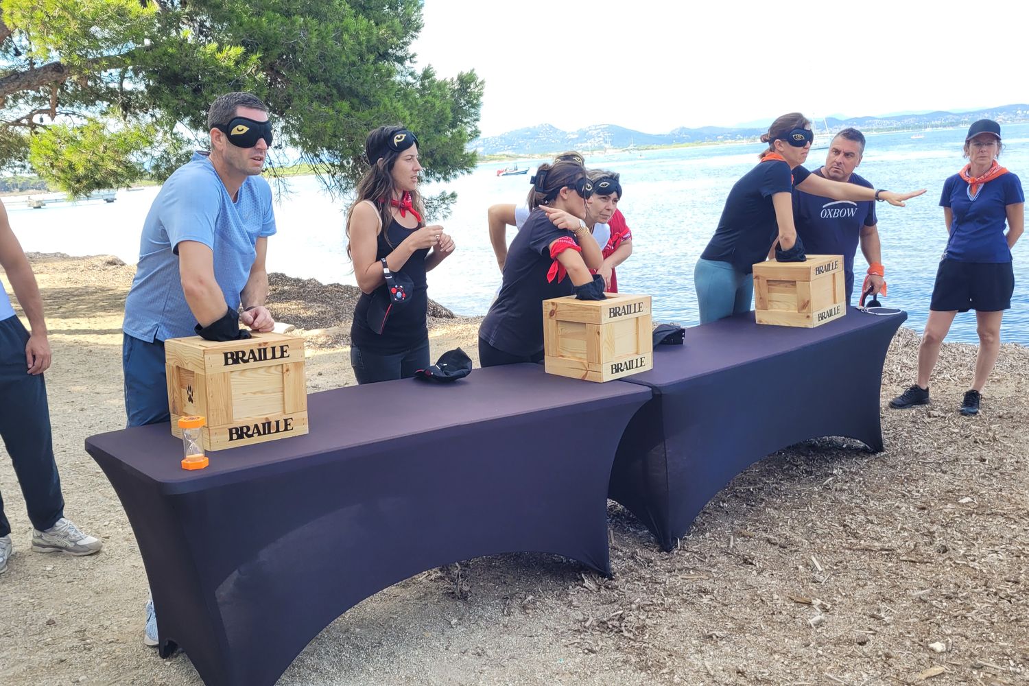 Équipes face à leurs caisses d'énigmes, sur une plage au bord de l'eau