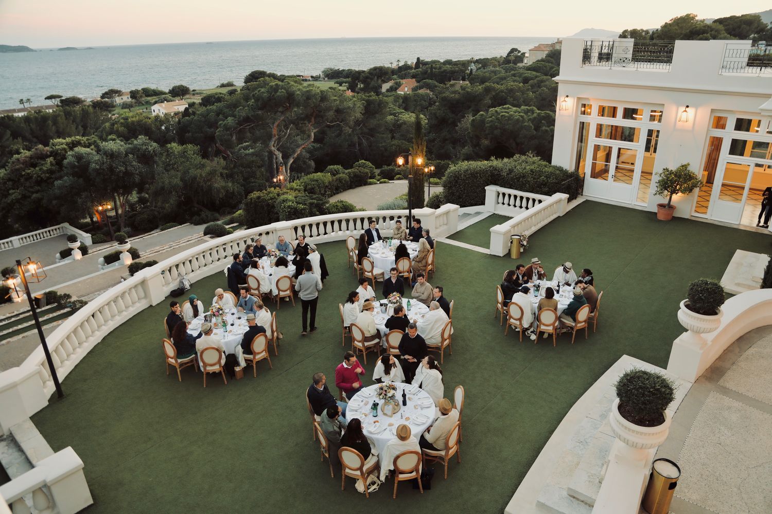 Dîner de gala sur la pelouse d'une villa face à la mer, tables rondes à la tombée du jour
