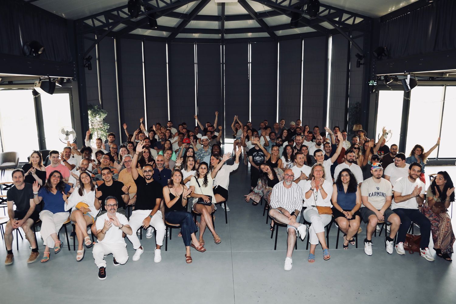 Salle plénière lumineuse remplie de participants assis, bras levés pour la photo