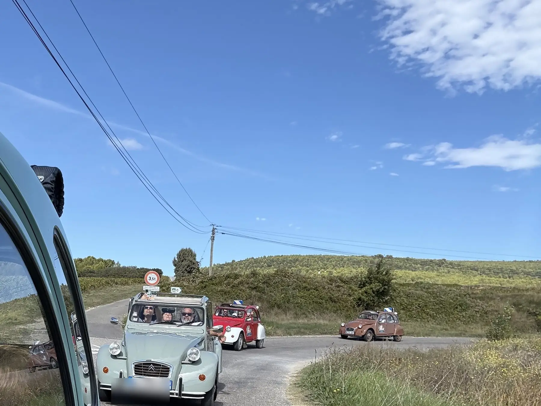 Convoi de 2 CV sur une route de l'arrière-pays, vu depuis une voiture